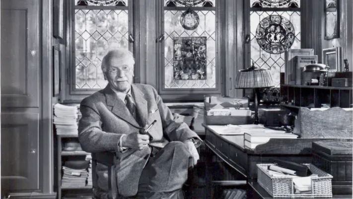 Carl Jung seated in his study, holding a pipe, surrounded by books and stained-glass windows